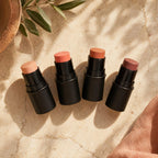 Four makeup sticks with different shades on a textured surface with a plant in the corner.