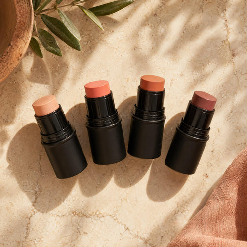 Four makeup sticks with different shades on a textured surface with a plant in the corner.