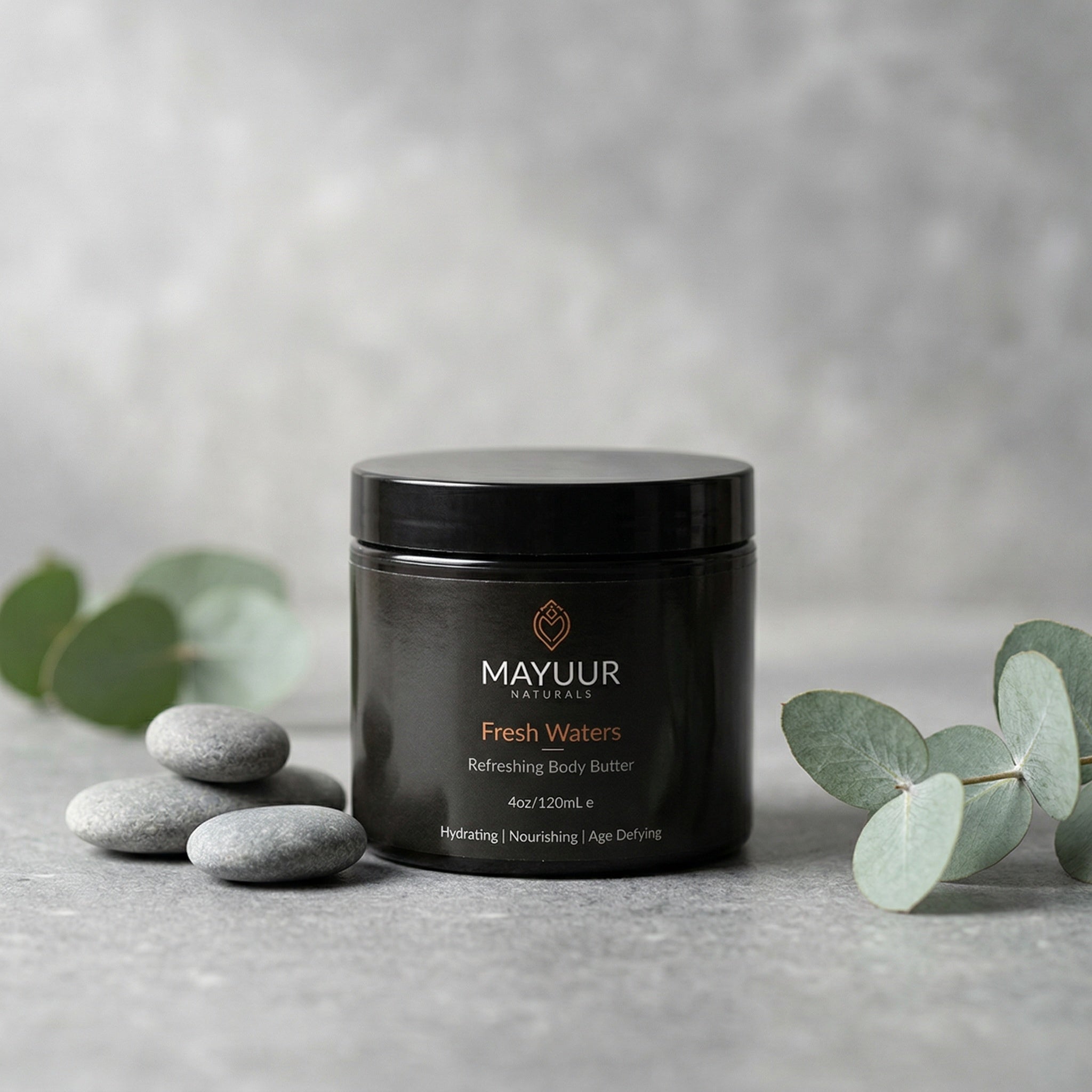 Black jar of Mayuur Fresh Waters body butter on a gray surface with eucalyptus leaves.