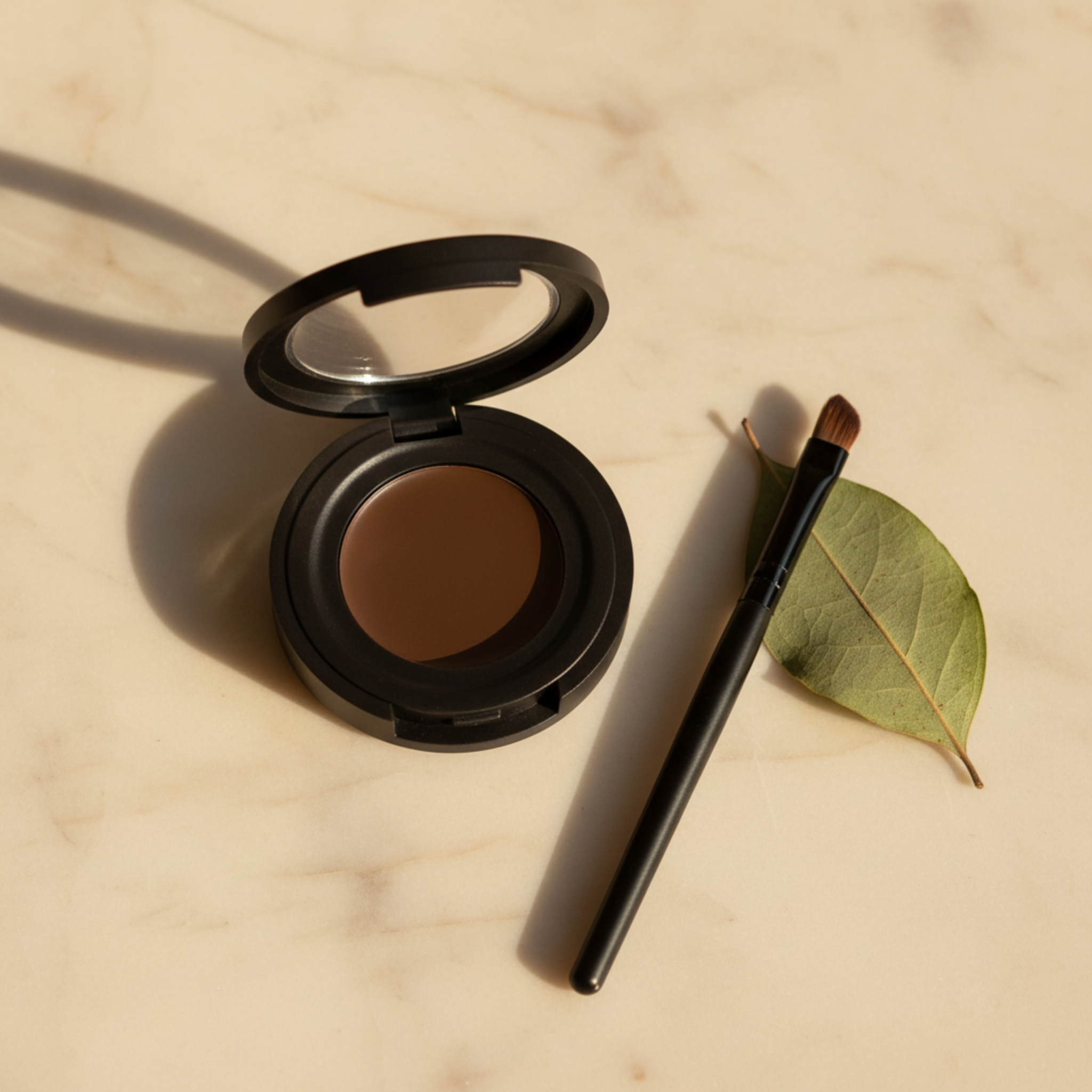 Open brown makeup compact with brush and leaf on beige surface