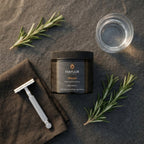 Mayuur Shaman body butter jar with a razor, towel, and rosemary on a dark surface