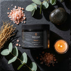 Mayuur Shaman Himalayan Salt Scrub on a dark surface with eucalyptus leaves and a candle.