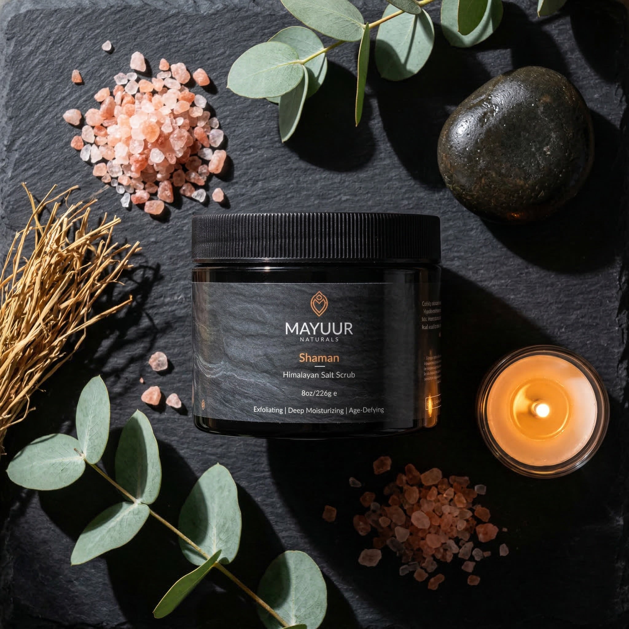 Mayuur Shaman Himalayan Salt Scrub on a dark surface with eucalyptus leaves and a candle.
