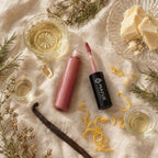 Two lip glosses on a textured surface with glasses of liquid, vanilla beans, and flowers.