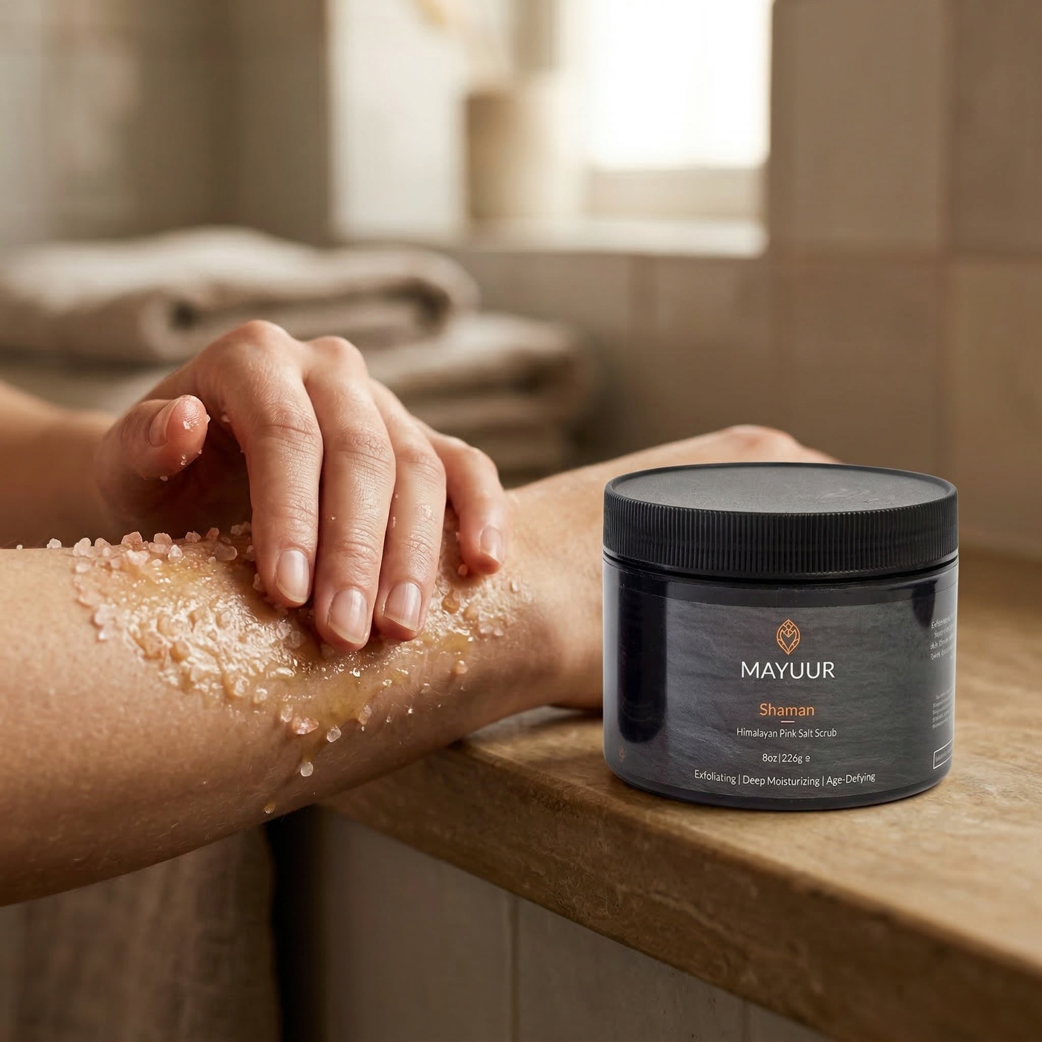 Person applying a exfoliating scrub to their arm with a jar of Mayuur Shaman scrub on a wooden surface.